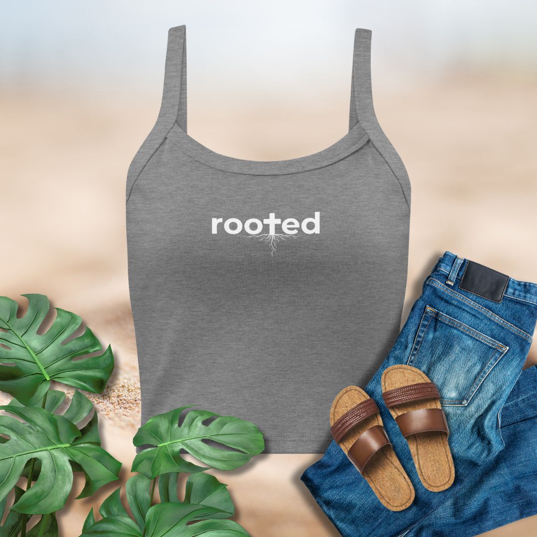 Gray tank top with 'rooted' text, blue jeans, brown sandals, and green leaves on a beige background