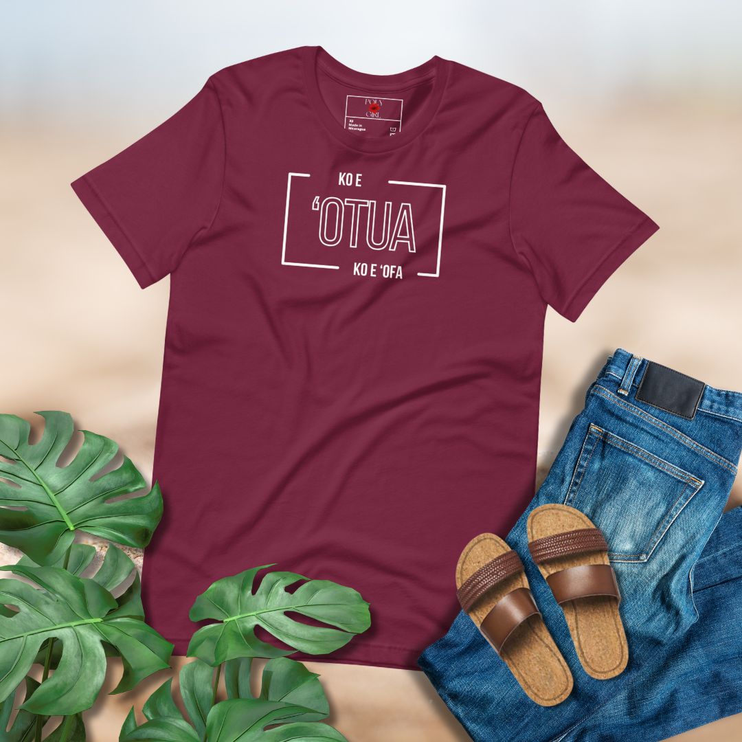 Maroon t-shirt with text "Ko e 'Otua Ko e 'Ofa" printed on front. Paired with sandals, jeans, and green leaves on a light background