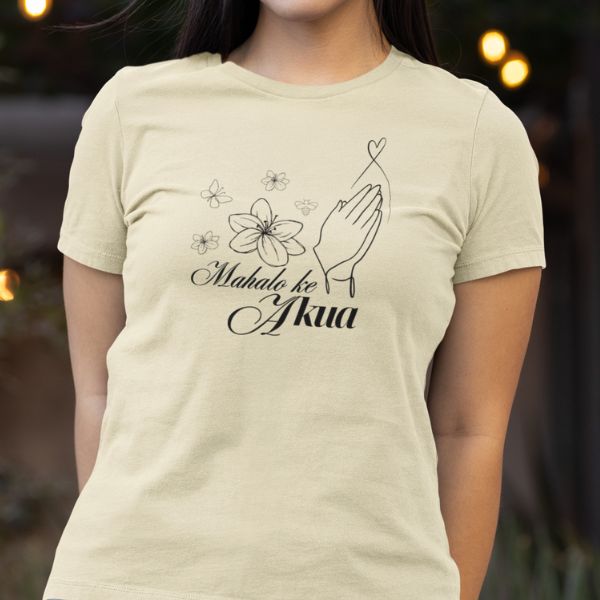 A woman wearing a beige t-shirt with the text 'Mahalo Ke Akua' and a graphic of hands and flowers on it.