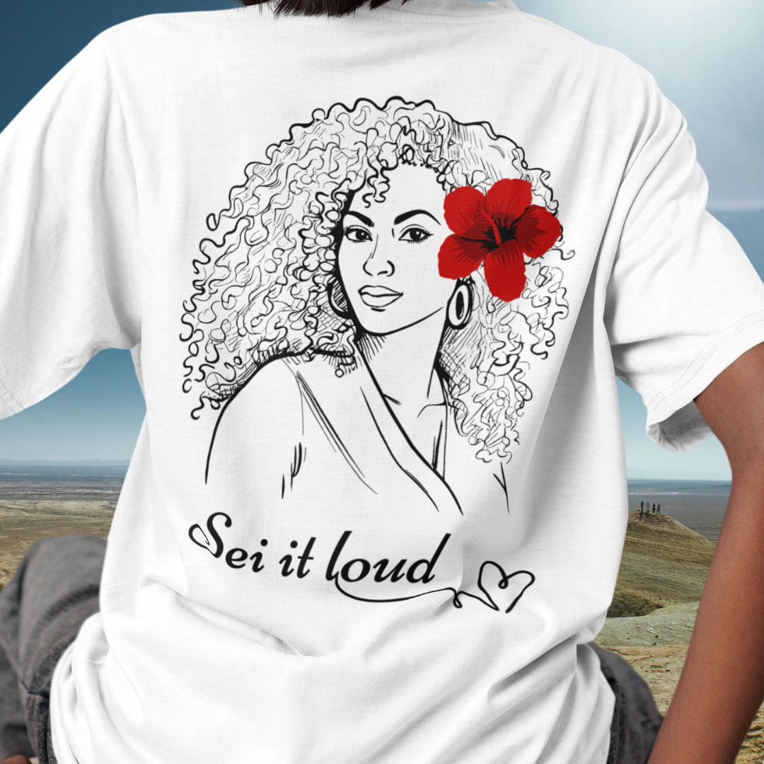 Woman wearing a white t-shirt with a design on the back of a woman with a red hibiscus in her hair and the word "Sei it loud".