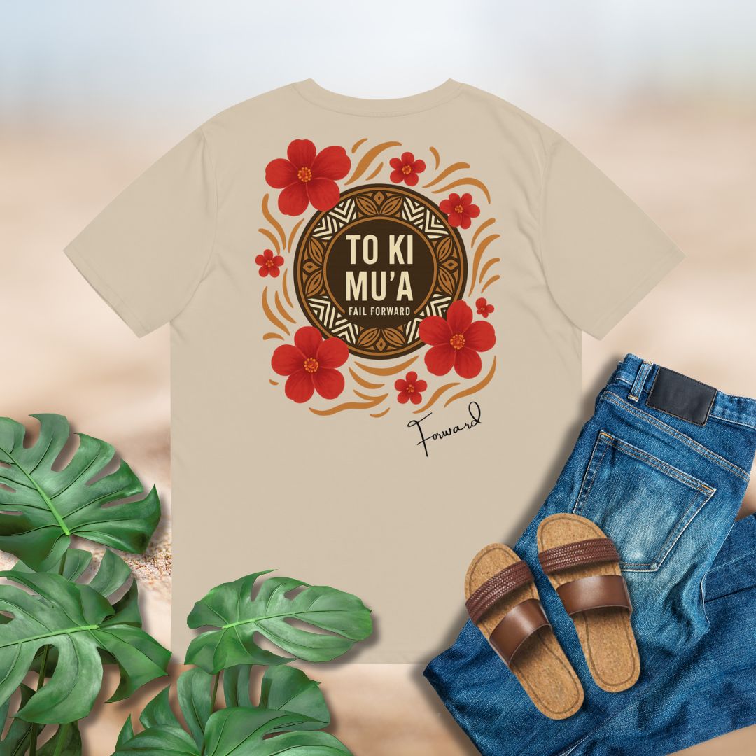 Beige t-shirt with a decorative design featuring red flowers and text of To Ki Mu'a on back of shirt placed on flat surface with jeans and sandals.