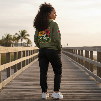 Woman wearing a green jacket with floral designs on a wooden boardwalk by the beach. Aga'i i luma design by Poly Girl.