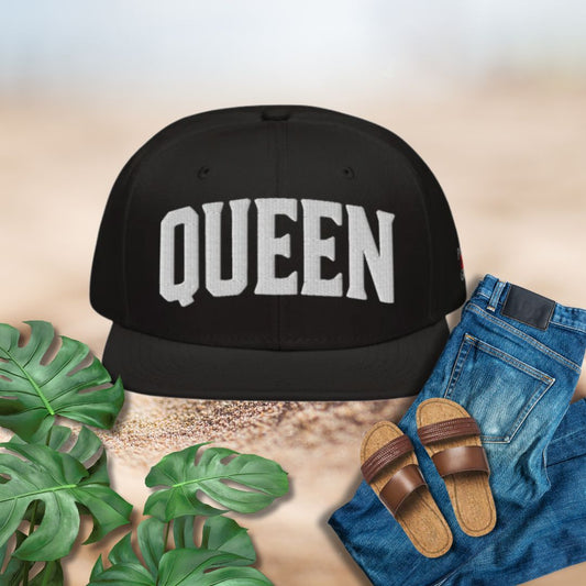 Black cap with 'QUEEN' text, sandals, and jeans on a textured surface with plants.