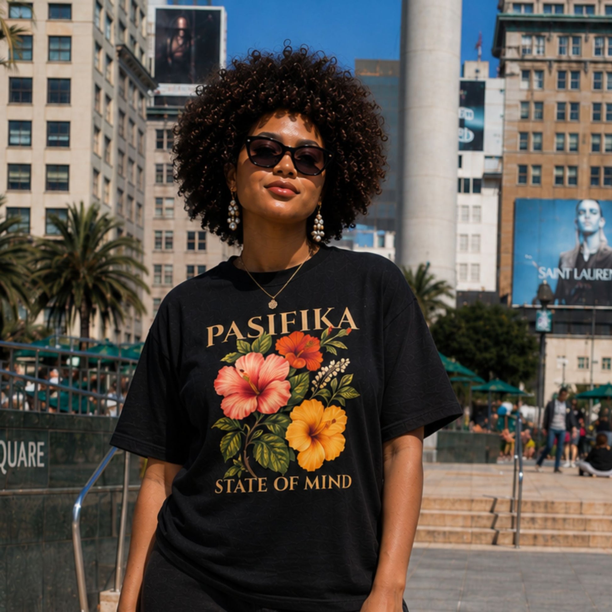 A woman wearing a black graphic t-shirt with a tropical floral print and the text 'PASIFIKA STATE OF MIND' stands outdoors in a city plaza with buildings and palm trees in the background.