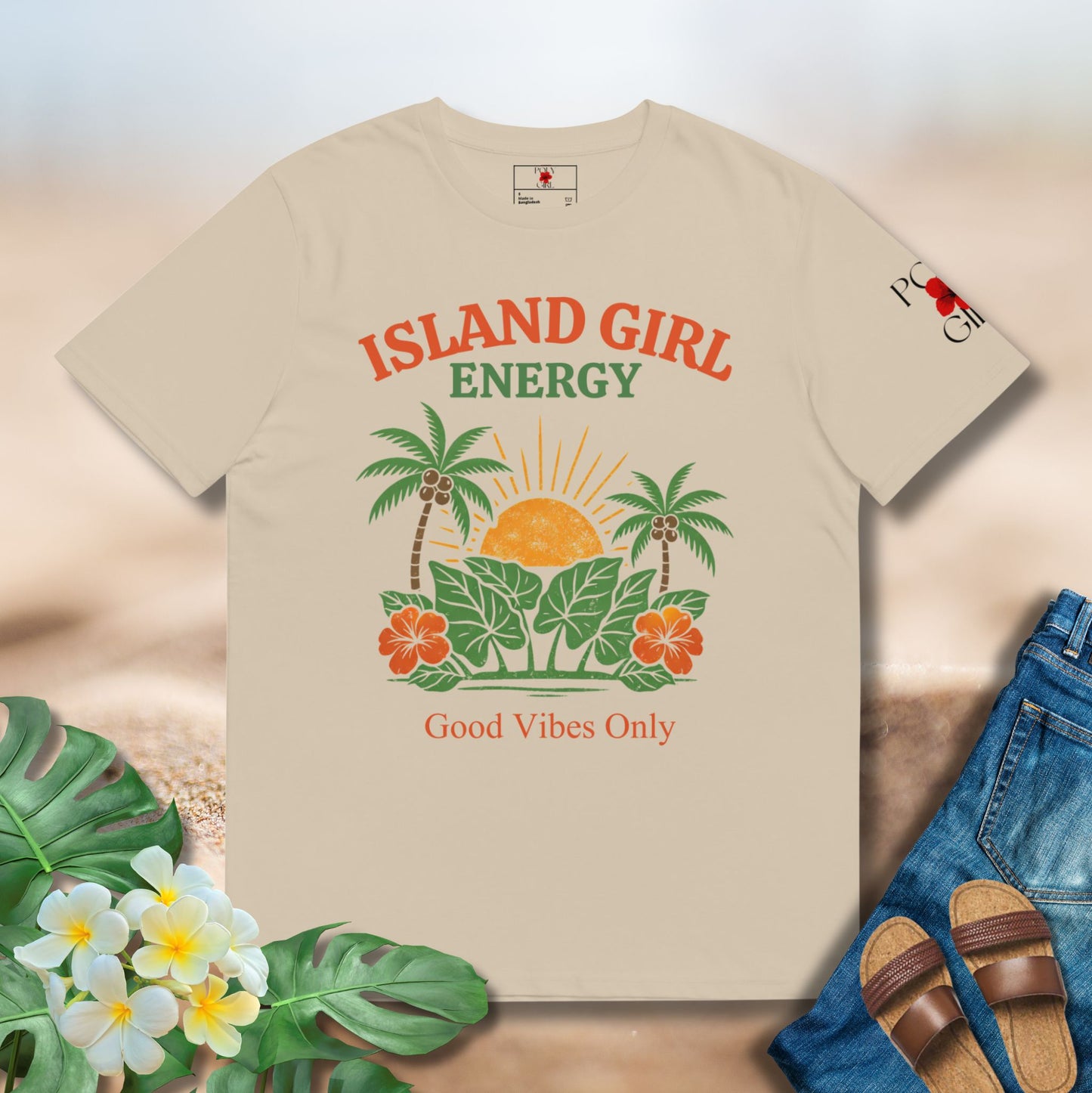 Beige t-shirt with 'Island Girl Energy' design on a sandy background with flowers and sandals.
