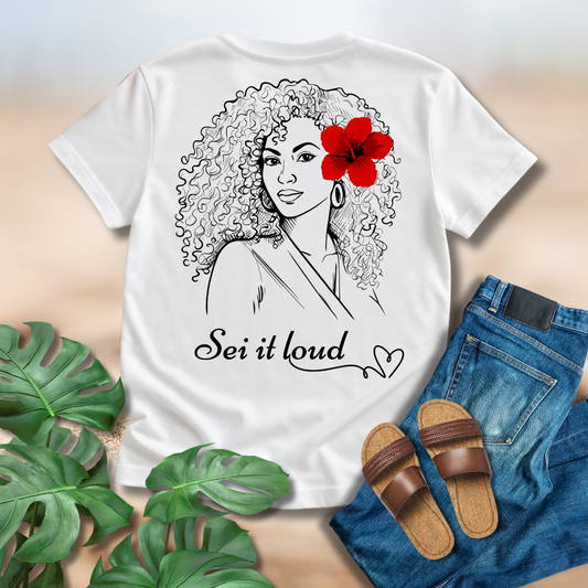 White t-shirt with a graphic of a woman with a red hibiscus in her hair and 'Sei it loud' text, surrounded by jeans, sandals, and plants.