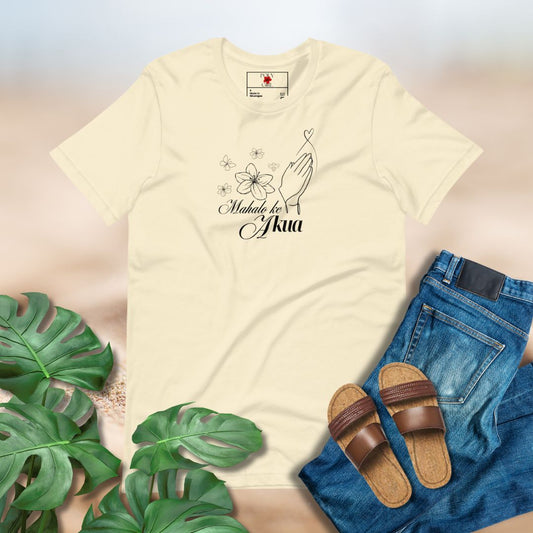 Yellow tshirt with Mahalo Ke Akua written on the front with prayer hands, flowers and butterflies paired with blue jeans, sandals and tropical leaves against a plain backdrop. 