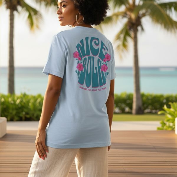 A Fijian Woman wearing a light blue t-shirt with Nice Bola text, standing on a wooden deck with palm trees and ocean view.