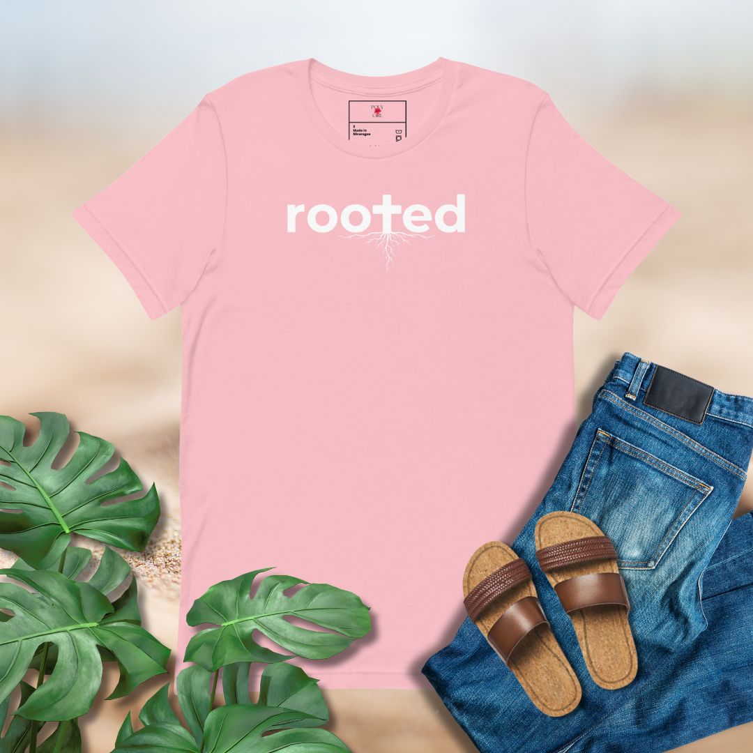 Pink t-shirt with 'rooted' text, paired with jeans, sandals, and plants on a neutral background