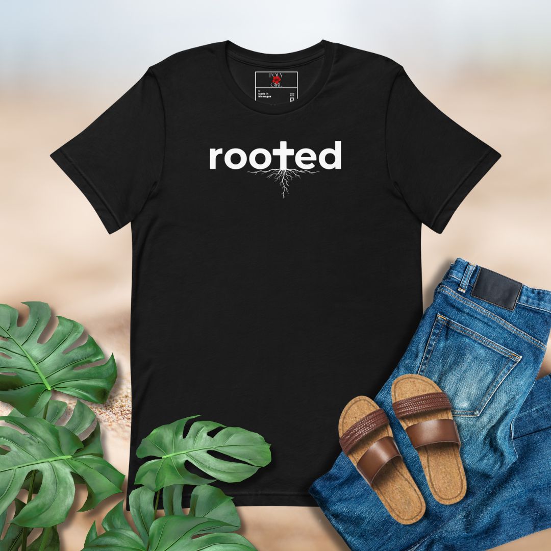 Black t-shirt with 'rooted' text, paired with jeans, sandals, and green leaves on a neutral background