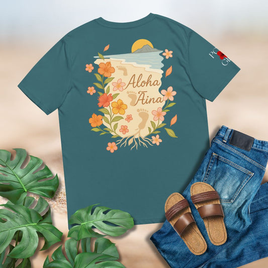 Teal t-shirt with floral design and 'Aloha Aina' text, surrounded by green leaves, sandals, and jeans on a light brown background.