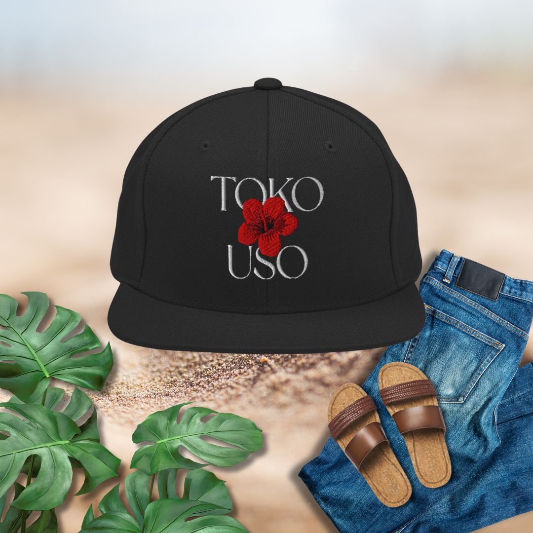 Black cap with 'TOKO USO' and a red flower design, paired with jeans and sandals on a neutral background.